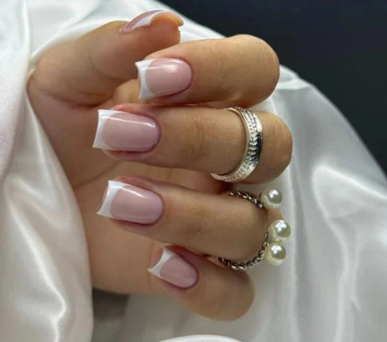 French Pink Silcare builder gel manicure with glossy pink base and white French tips on square-shaped nails, shown on a hand wearing silver rings and pearl jewellery against silky white fabric background, highlighting elegant salon-style nails.