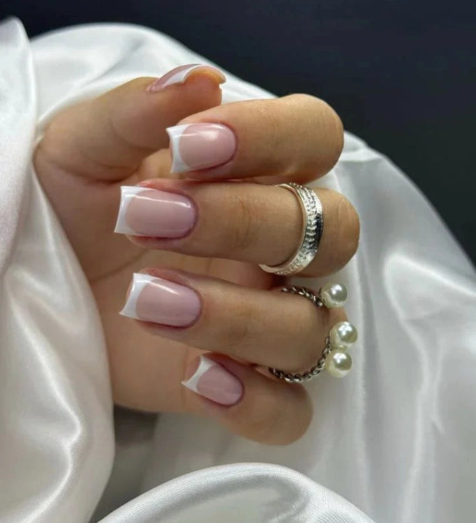 French Pink Silcare builder gel manicure with glossy pink base and white French tips on square-shaped nails, shown on a hand wearing silver rings and pearl jewellery against silky white fabric background, highlighting elegant salon-style nails.