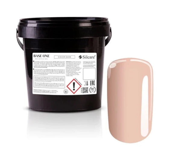 Silcare Base One Cover Dark Builder UV Gel in a black bulk container with the product label visible, displayed next to a glossy nude beige nail swatch showing the camouflage cover shade. The image highlights professional salon-size builder gel packaging designed for nail extensions, nail bed enhancement, overlays, and nail sculpting.